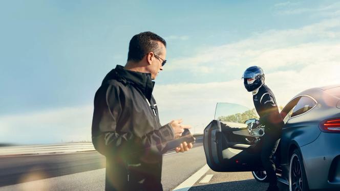 A man holding a phone greets a helmed driver leaving an AMG vehicle