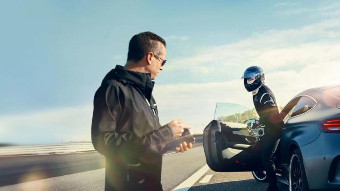 A man holding a phone greets a helmed driver leaving an AMG vehicle