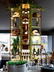 In einem von grünen Ranken umschlugenen, goldfarbenem Regal liegen verschiedene Helme, Gläser und Champagnerflaschen.