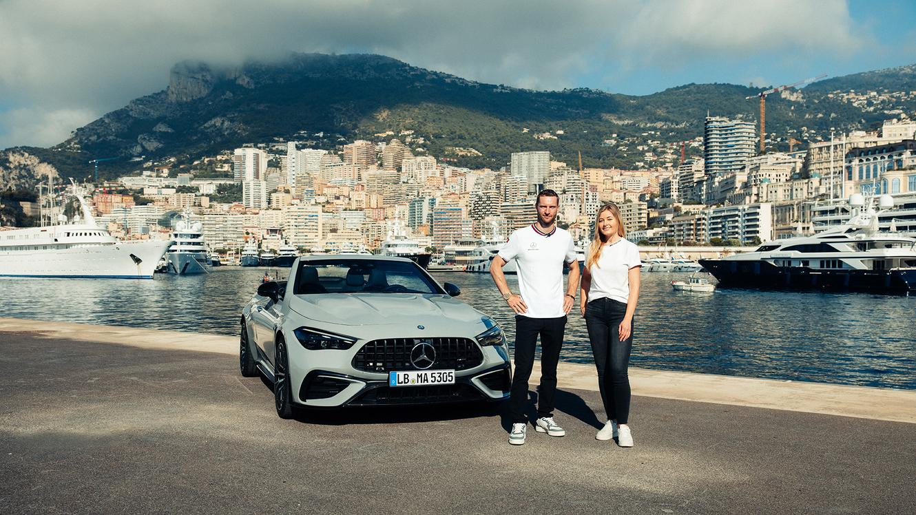 Maro Engel und Sabrine Berchtold posieren mit dem neuen Mercedes-AMG CLE 53 4MATIC+ Cabriolet vor einer Hafenstadt