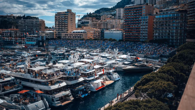 Im Hafenbecken von Monaco stehen diverse Yachten nebeneinander während im Hintergrund die vollen Tribünen an der Formel 1 Strecke zu sehen sind.