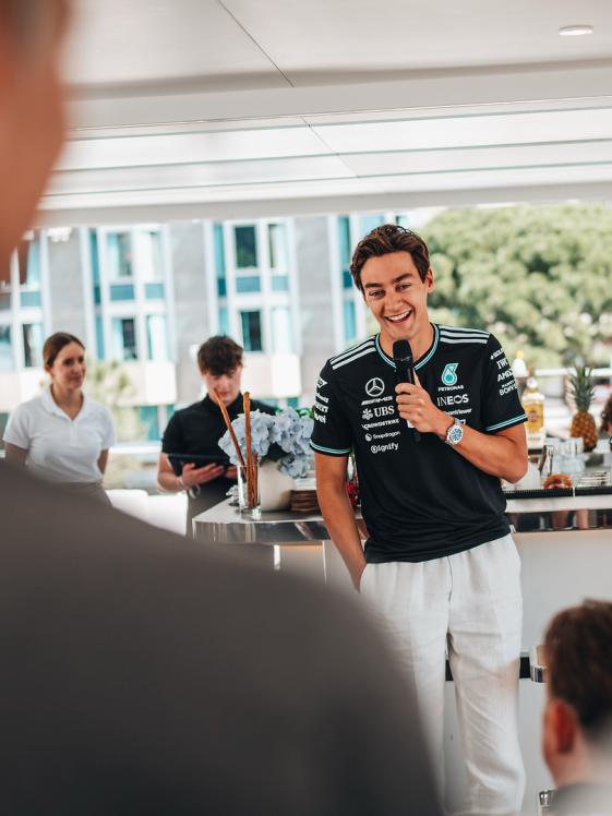 George Russell steht lachend mit einem Mikrofon in der Hand vor einem Bartresen.